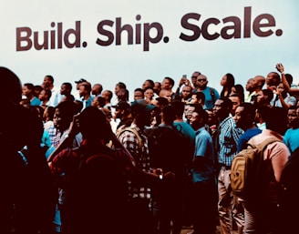 A large group of people are gathered together, many of them holding or using smartphones, suggesting an event or gathering. The backdrop features the words 'Build. Ship. Scale.' in large bold letters, indicating a tech or entrepreneurial theme. The crowd consists of individuals dressed casually and engaging with each other, capturing a sense of community and interaction.