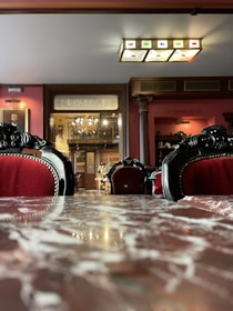 An elegant, classic interior with plush red velvet chairs and intricate black detailing. A reflective marble table is in the foreground. The room features wooden paneling, column details, and a decorative ceiling light. Portraits and antiques adorn the warm-toned walls.