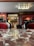 An elegant, classic interior with plush red velvet chairs and intricate black detailing. A reflective marble table is in the foreground. The room features wooden paneling, column details, and a decorative ceiling light. Portraits and antiques adorn the warm-toned walls.