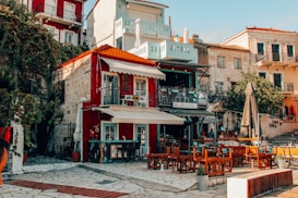 A vibrant outdoor cafe with wooden furniture is set against a backdrop of colorful, multi-story buildings. The walls of the buildings showcase a mix of red, white, and pale yellow hues, with balconies, plants, and various architectural details.