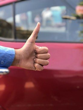 A hand giving a thumbs-up gesture in front of a red car. The person is wearing a blue shirt. The background is slightly out of focus with the car door visible.