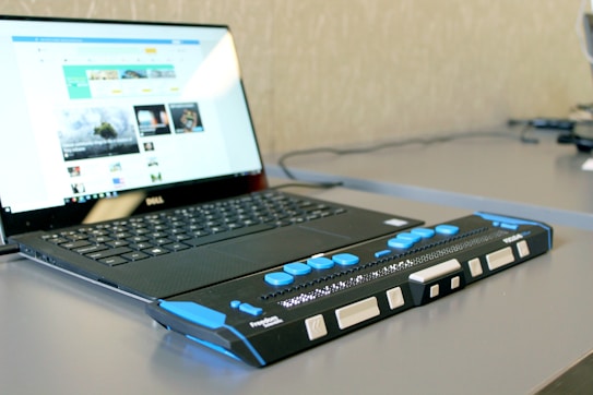 A laptop with an open screen displaying a web page is situated on a desk. In front of the laptop is a Braille display device, featuring tactile buttons and controls, designed to assist visually impaired users in reading digital content.