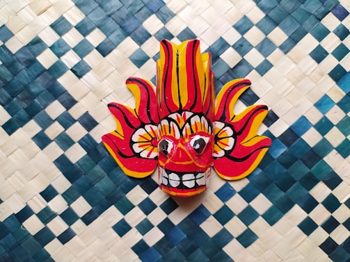 A vibrant, colorful mask with an expression of energy and intensity, featuring red and yellow flames extending from the top and sides. The mask is set against a woven pattern background of blue and white squares.