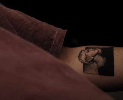 A detailed black and gray tattoo featuring a side profile of a person's head, with short hair and a solemn expression. The tattoo is located on a person's forearm, and the person is wearing a muted brown shirt. The background is dark, adding contrast to the tattoo's intricate detail.