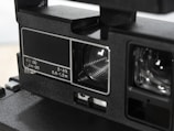 Close-up of a vintage camera component featuring detailed markings in white, indicating focus distances. The focus area and lens are visible, captured with a sleek, matte black finish. The image highlights the mechanical and optical elements of the camera's design.