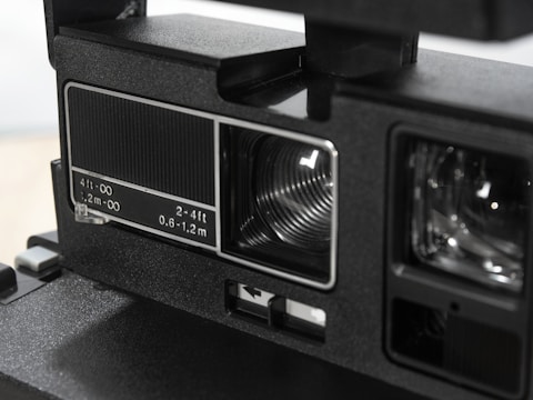 Close-up of a vintage camera component featuring detailed markings in white, indicating focus distances. The focus area and lens are visible, captured with a sleek, matte black finish. The image highlights the mechanical and optical elements of the camera's design.