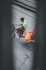 A person is riding a rickshaw on a street, viewed from above. The rickshaw has an orange seat and the rider is wearing a bright green vest with writing on the back. The road is gray, and there are black cables running across the top of the image.
