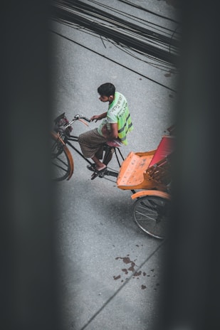 A person is riding a rickshaw on a street, viewed from above. The rickshaw has an orange seat and the rider is wearing a bright green vest with writing on the back. The road is gray, and there are black cables running across the top of the image.