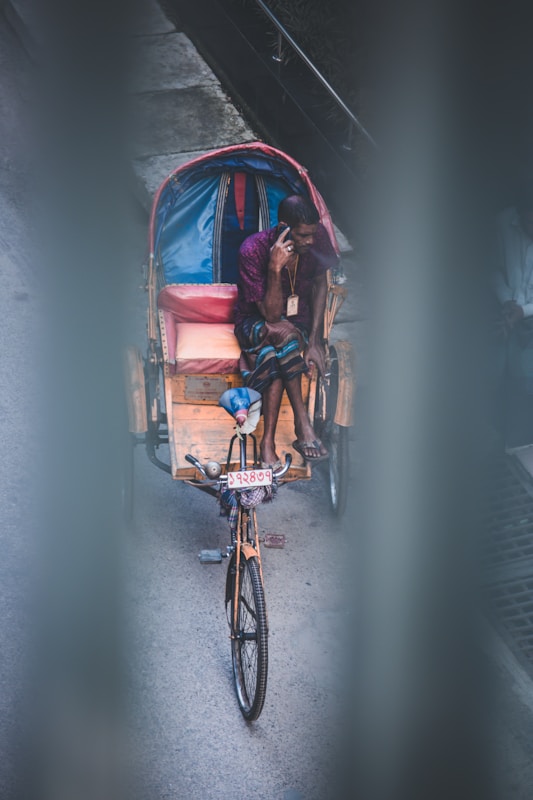 A person is seated on a bicycle rickshaw, talking on a mobile phone. The rickshaw has a blue and pink interior with a number plate visible at the front. The scene is viewed from above, through blurred vertical bars.