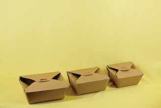 Three brown cardboard takeout boxes are neatly arranged in a row against a light yellow background. The boxes are closed with folded flaps on the top.