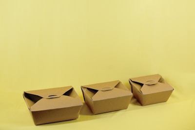 Three brown cardboard takeout boxes are neatly arranged in a row against a light yellow background. The boxes are closed with folded flaps on the top.