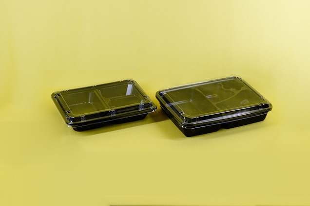 Two black plastic food containers with transparent lids are positioned on a solid yellow surface. The containers have separate compartments inside, designed for holding different types of food.