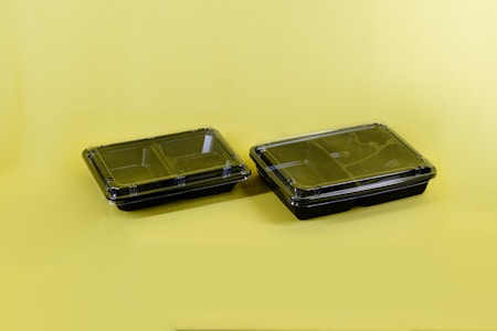 Two black plastic food containers with transparent lids are positioned on a solid yellow surface. The containers have separate compartments inside, designed for holding different types of food.
