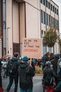 A group of people are gathered outdoors, participating in a protest or demonstration. The focus is on a person holding a cardboard sign that reads 'We are the kids, we make the difference.' The group seems to be walking along a street, surrounded by buildings and trees. The atmosphere is active and engaged, with participants wearing warm clothing.
