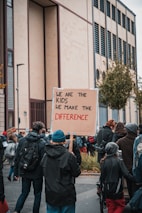 A group of people are gathered outdoors, participating in a protest or demonstration. The focus is on a person holding a cardboard sign that reads 'We are the kids, we make the difference.' The group seems to be walking along a street, surrounded by buildings and trees. The atmosphere is active and engaged, with participants wearing warm clothing.