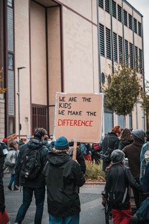 A group of people are gathered outdoors, participating in a protest or demonstration. The focus is on a person holding a cardboard sign that reads 'We are the kids, we make the difference.' The group seems to be walking along a street, surrounded by buildings and trees. The atmosphere is active and engaged, with participants wearing warm clothing.