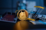 A golden coin featuring a cartoon dog is prominently placed in the center, surrounded by electronic components like circuit boards and resistors. The focus is sharp on the coin, while the background is blurred, giving a sense of depth.