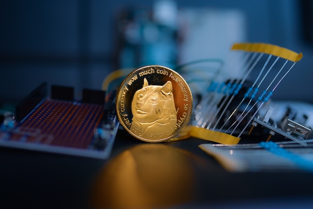 A golden coin featuring a cartoon dog is prominently placed in the center, surrounded by electronic components like circuit boards and resistors. The focus is sharp on the coin, while the background is blurred, giving a sense of depth.