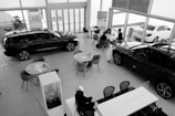 A car dealership showroom featuring several tables and chairs arranged for consultations or meetings. Two cars are prominently displayed indoors, while another is visible through the large glass windows. There are people engaged in discussions at one of the tables.