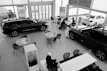 A car dealership showroom featuring several tables and chairs arranged for consultations or meetings. Two cars are prominently displayed indoors, while another is visible through the large glass windows. There are people engaged in discussions at one of the tables.