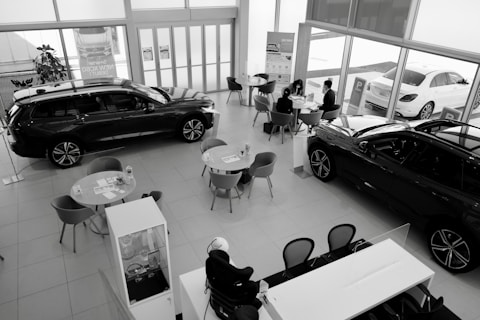 A car dealership showroom featuring several tables and chairs arranged for consultations or meetings. Two cars are prominently displayed indoors, while another is visible through the large glass windows. There are people engaged in discussions at one of the tables.