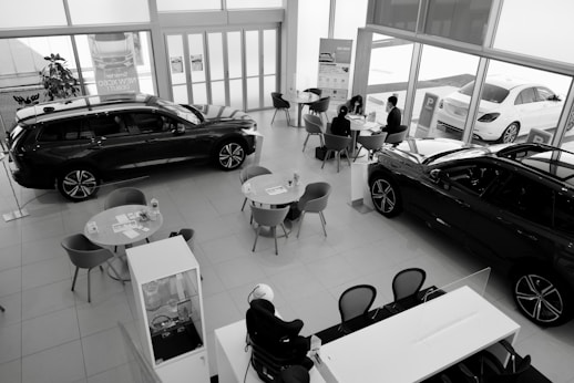 A car dealership showroom featuring several tables and chairs arranged for consultations or meetings. Two cars are prominently displayed indoors, while another is visible through the large glass windows. There are people engaged in discussions at one of the tables.
