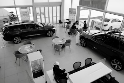 A car dealership showroom featuring several tables and chairs arranged for consultations or meetings. Two cars are prominently displayed indoors, while another is visible through the large glass windows. There are people engaged in discussions at one of the tables.