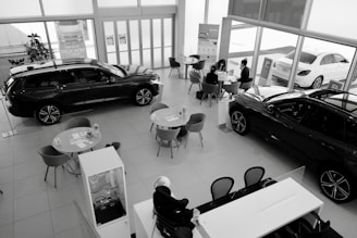 A car dealership showroom featuring several tables and chairs arranged for consultations or meetings. Two cars are prominently displayed indoors, while another is visible through the large glass windows. There are people engaged in discussions at one of the tables.