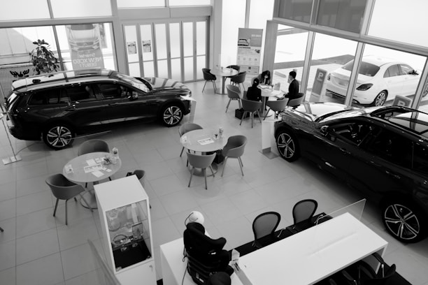 A car dealership showroom featuring several tables and chairs arranged for consultations or meetings. Two cars are prominently displayed indoors, while another is visible through the large glass windows. There are people engaged in discussions at one of the tables.