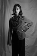 A black and white portrait of a person with short hair standing with one hand on their hip. The person is wearing a long-sleeved shirt and trousers. The background features a textured fabric backdrop, creating a studio setting.
