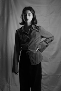 A black and white portrait of a person with short hair standing with one hand on their hip. The person is wearing a long-sleeved shirt and trousers. The background features a textured fabric backdrop, creating a studio setting.
