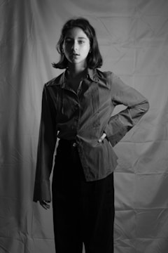 A black and white portrait of a person with short hair standing with one hand on their hip. The person is wearing a long-sleeved shirt and trousers. The background features a textured fabric backdrop, creating a studio setting.