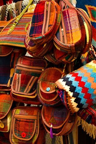 A collection of colorful, woven bags with intricate patterns, featuring various shades of red, orange, and blue. The bags have button closures and are arranged in a layered display, giving a vibrant and artistic impression.