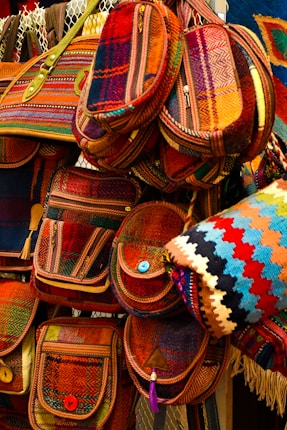 A collection of colorful, woven bags with intricate patterns, featuring various shades of red, orange, and blue. The bags have button closures and are arranged in a layered display, giving a vibrant and artistic impression.