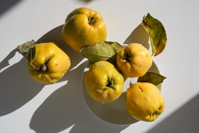 Five yellow quinces with rough skins are placed on a white surface, accompanied by a few green leaves. The light creates distinct shadows, enhancing the texture and color of the fruit.