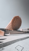 A modern architectural structure with a striking, elongated, and layered design resembling a stack of wooden beams. The building appears to be a pavilion or exhibition hall, with a light-colored facade and people walking around the premises. The background includes a clear sky and adjacent modern buildings.