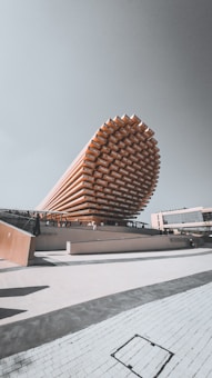 A modern architectural structure with a striking, elongated, and layered design resembling a stack of wooden beams. The building appears to be a pavilion or exhibition hall, with a light-colored facade and people walking around the premises. The background includes a clear sky and adjacent modern buildings.