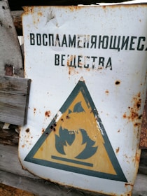 A weathered and rusted sign with a triangular warning symbol indicating flammable materials, surrounded by a wooden background. The text above the symbol is in a different language.
