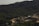 A dense forest with rich greenery covers the hilly landscape, while a mining or construction site with multiple mounds of earth and gravel is situated in the foreground. The site includes a few small buildings and unpaved roads, suggesting industrial activity amidst the natural surroundings.