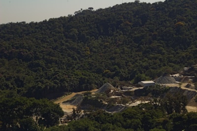 A dense forest with rich greenery covers the hilly landscape, while a mining or construction site with multiple mounds of earth and gravel is situated in the foreground. The site includes a few small buildings and unpaved roads, suggesting industrial activity amidst the natural surroundings.