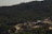A dense forest with rich greenery covers the hilly landscape, while a mining or construction site with multiple mounds of earth and gravel is situated in the foreground. The site includes a few small buildings and unpaved roads, suggesting industrial activity amidst the natural surroundings.