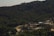 A dense forest with rich greenery covers the hilly landscape, while a mining or construction site with multiple mounds of earth and gravel is situated in the foreground. The site includes a few small buildings and unpaved roads, suggesting industrial activity amidst the natural surroundings.