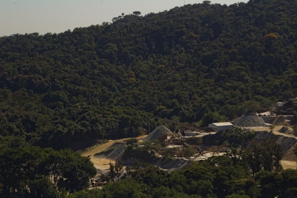 A dense forest with rich greenery covers the hilly landscape, while a mining or construction site with multiple mounds of earth and gravel is situated in the foreground. The site includes a few small buildings and unpaved roads, suggesting industrial activity amidst the natural surroundings.