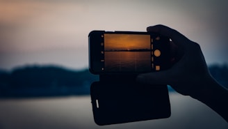 A silhouette of a hand holding a smartphone capturing a sunset over a calm body of water. The phone screen shows a warm, orange horizon with subtle reflections on the water. The background is dimly lit, adding contrast to the bright image on the phone display.