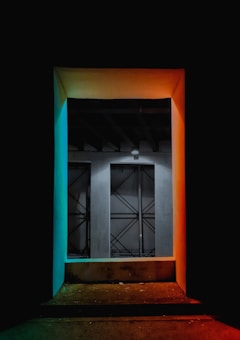 A doorway is highlighted by colored lights with a gradient of teal on the left and orange on the right. Inside, a gray wall with a geometric pattern is visible. The floor is dark and slightly reflective.