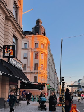 A vibrant city street scene with a sunset-lit building featuring an ornate dome and light brown and white facade. Below, a Kebab House sign is visible alongside other shops. Several people are walking, and a person pushes a stroller. Scooters are parked and traffic moves in the background under a clear blue sky.