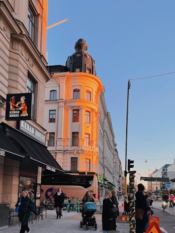 A vibrant city street scene with a sunset-lit building featuring an ornate dome and light brown and white facade. Below, a Kebab House sign is visible alongside other shops. Several people are walking, and a person pushes a stroller. Scooters are parked and traffic moves in the background under a clear blue sky.
