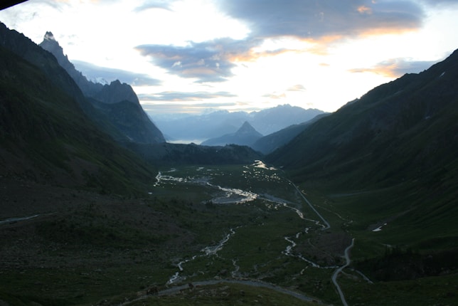 A picturesque valley with winding streams reflecting the light of a colorful sunset. Majestic mountains surround the valley, creating a serene and expansive landscape. The sky features a mix of clouds and breaks of sunlight.