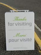 An outdoor sign displays a message in both English and French, expressing gratitude to visitors. The sign reads 'Thanks for visiting' and 'Merci pour visité' in green and gray text. It is located on a paved surface with a yellow painted curb visible in the background.