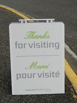 An outdoor sign displays a message in both English and French, expressing gratitude to visitors. The sign reads 'Thanks for visiting' and 'Merci pour visité' in green and gray text. It is located on a paved surface with a yellow painted curb visible in the background.